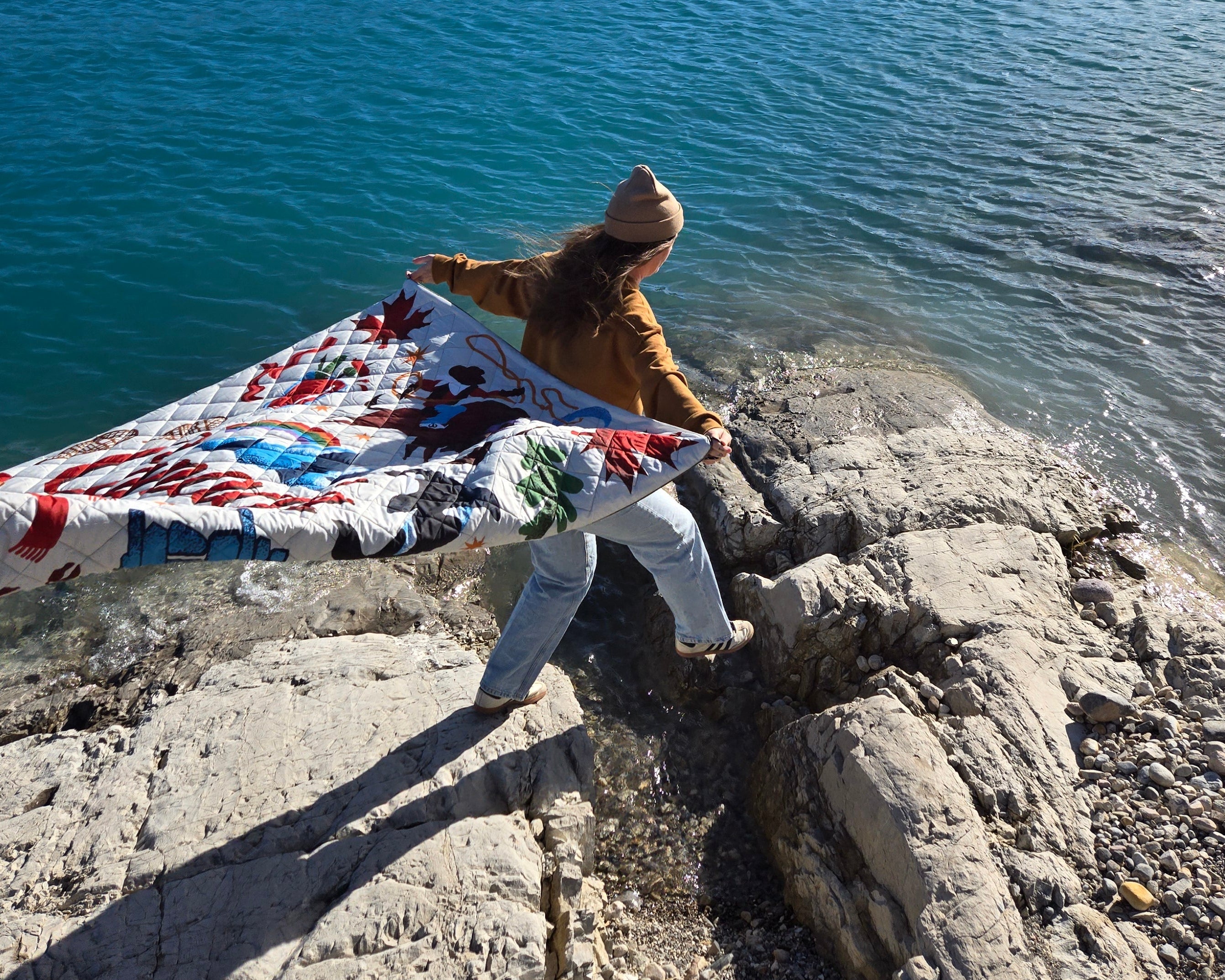 A person stands on a rocky lakeshore holding a colorful patterned blanket or quilt outdoors, with a large blue lake, forested island, and mountains in the background.