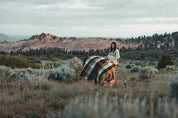 Woman holding cozy plaid blanket in scenic outdoor landscape with mountains and grass