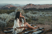 Woman relaxing outdoors on striped blanket with wine, surrounded by scenic desert landscape