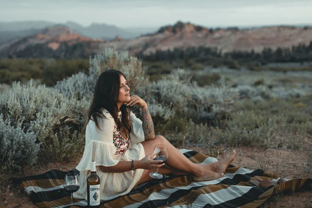 Woman relaxing outdoors on striped blanket with wine, surrounded by scenic desert landscape