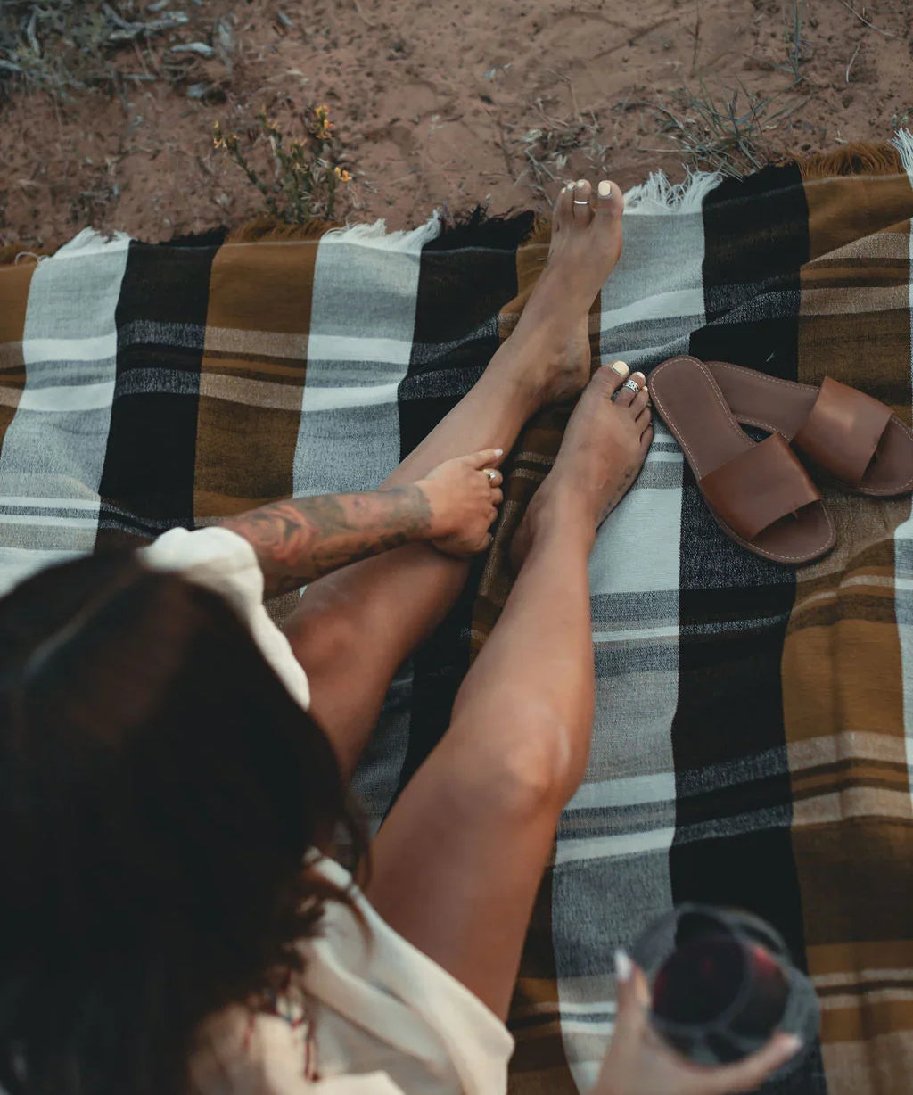 Woman relaxing outdoors on a plaid blanket with brown sandals and a drink