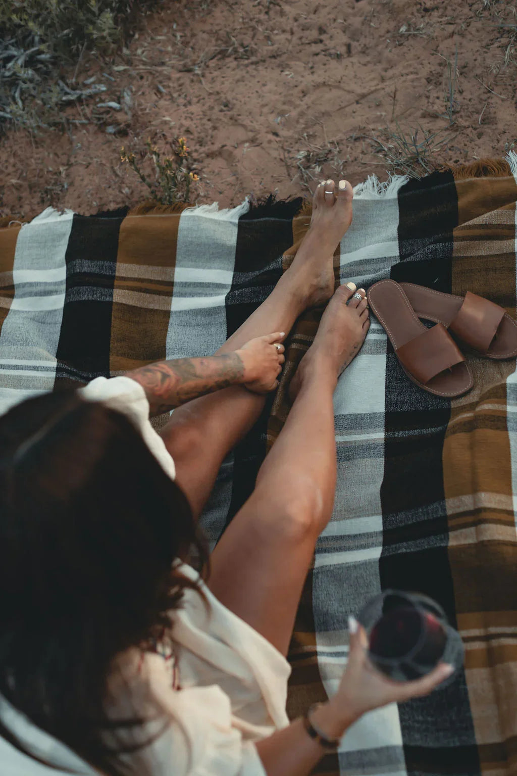 Woman relaxing outdoors on a plaid blanket with brown sandals and a drink