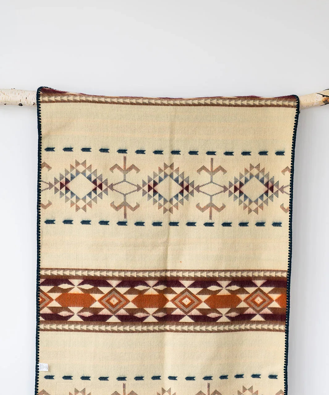 Beige woven blanket with geometric diamond patterns and brown, orange, and navy accents, hanging on a branch.