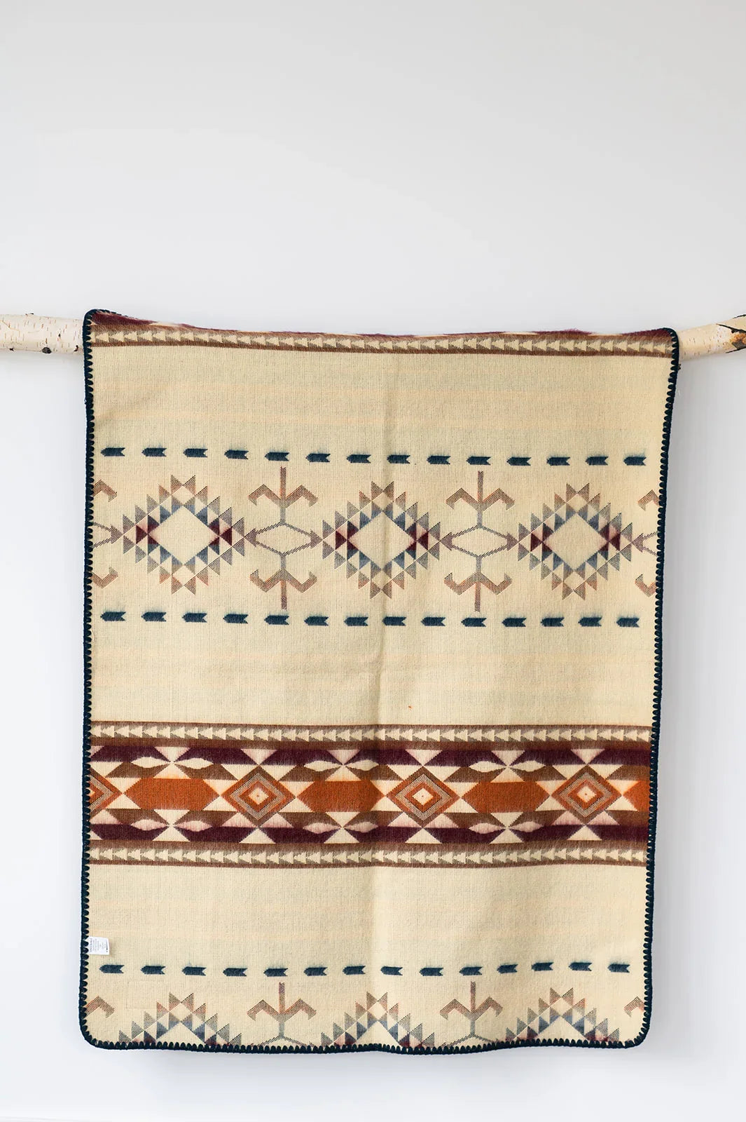 Beige woven blanket with geometric diamond patterns and brown, orange, and navy accents, hanging on a branch.