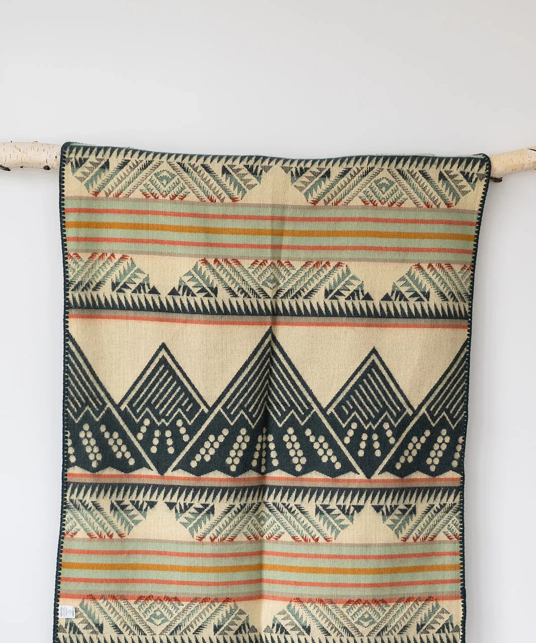 Beige woven blanket with geometric mountain and stripe pattern, hanging on a white wall