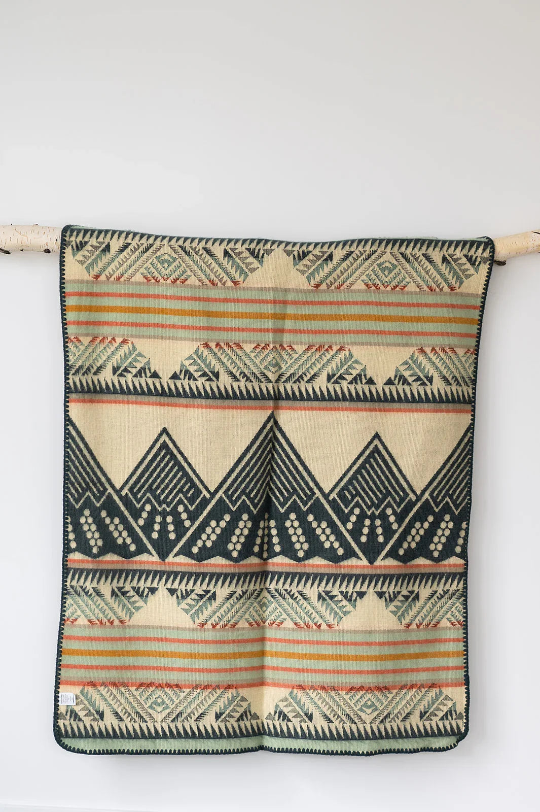 Beige woven blanket with geometric mountain and stripe pattern, hanging on a white wall