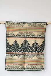 Beige woven blanket with geometric mountain and stripe pattern, hanging on a white wall