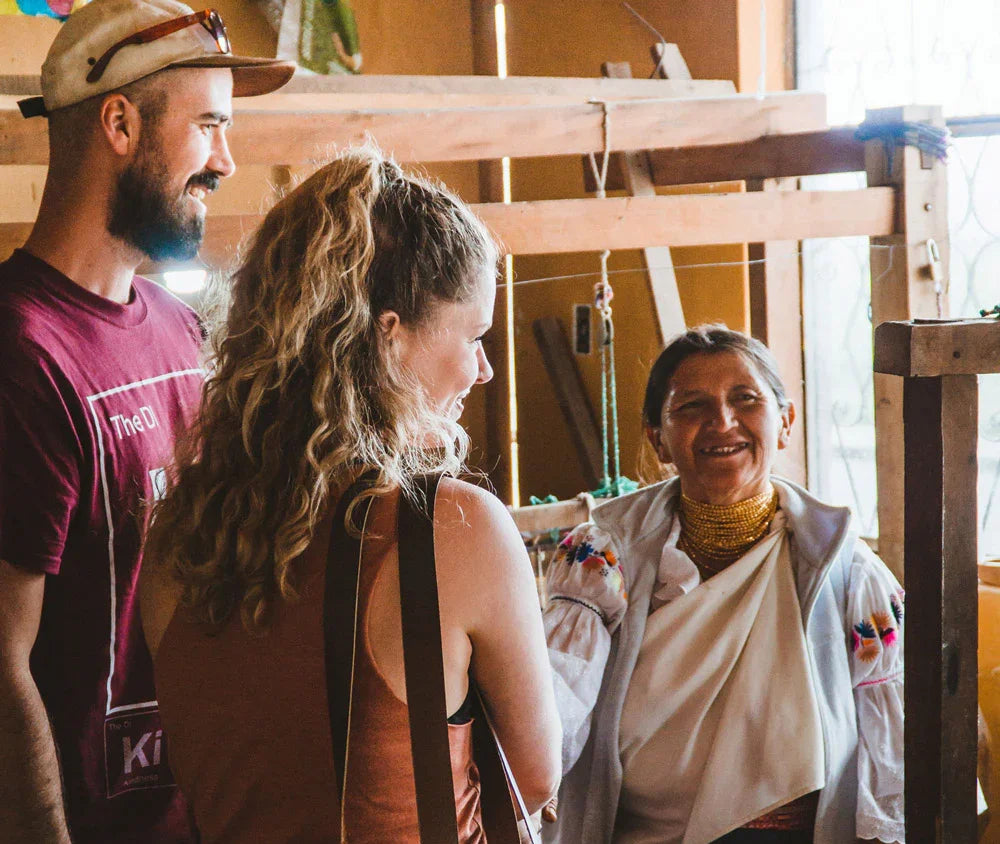 Three people smiling and talking in a cozy artisan workshop with wooden loom in background.