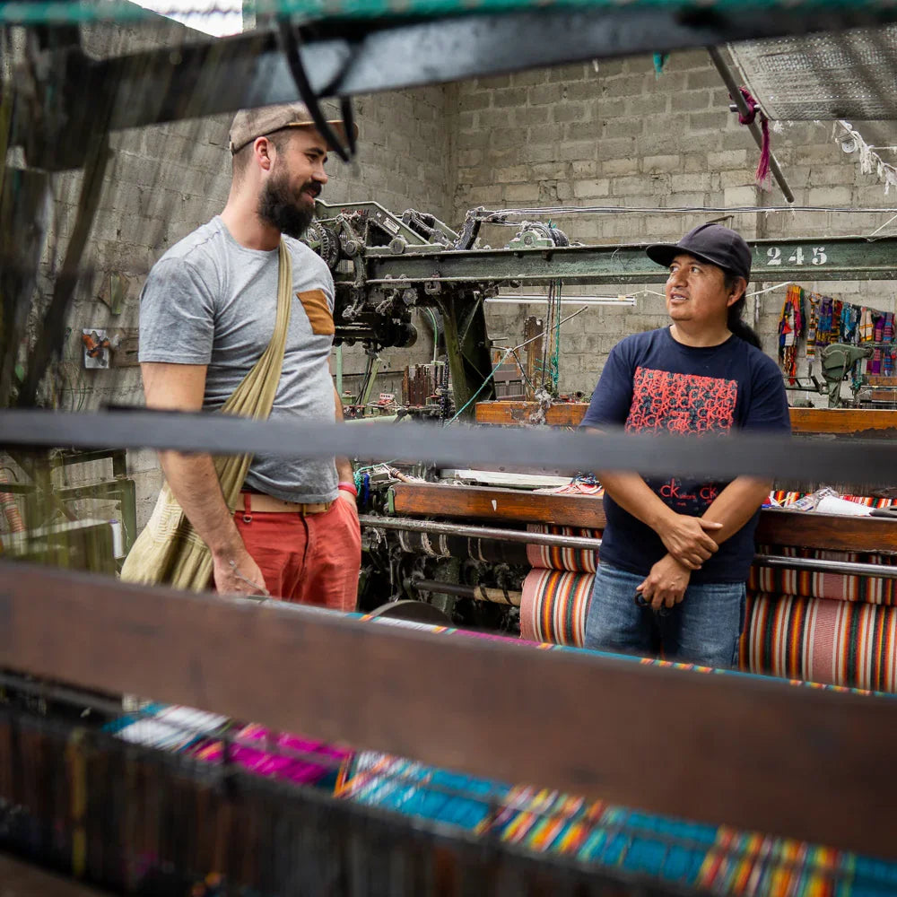 Two artisans talking in a textile workshop with colorful woven blankets and looms