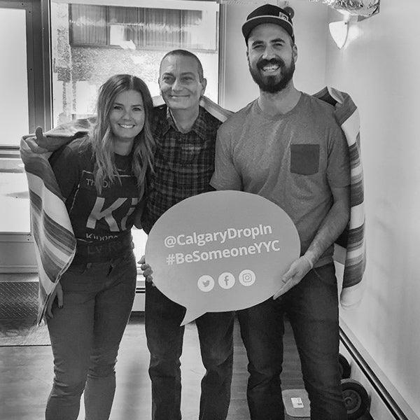 Three people smiling indoors, two sharing a blanket, holding a Calgary Drop-In sign promoting community support.