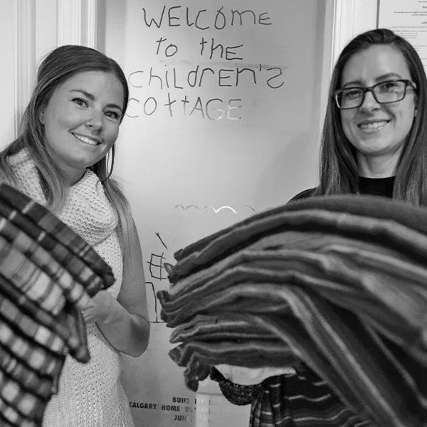 Two women smiling and holding stacks of blankets in front of a door labeled Children's Cottage