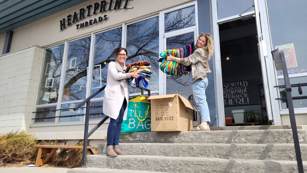 Two women holding colorful blankets outside a storefront, donation boxes on steps