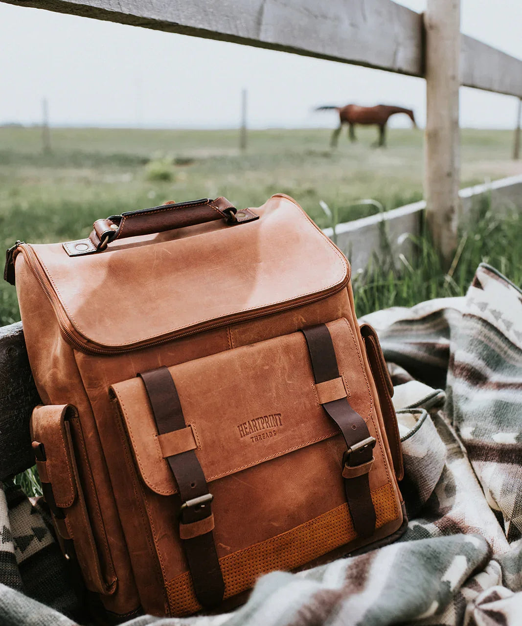 Brown leather backpack on patterned blanket outdoors by a fence, horse in background, rustic setting