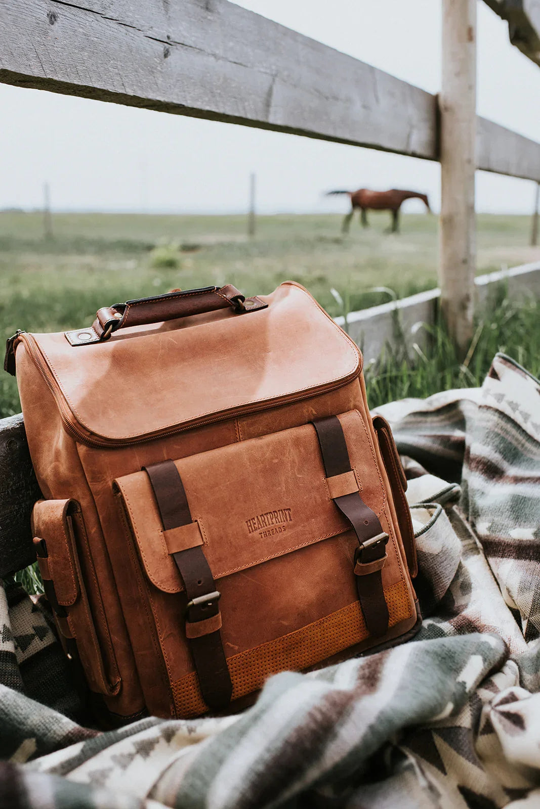 Brown leather backpack on patterned blanket outdoors by a fence, horse in background, rustic setting