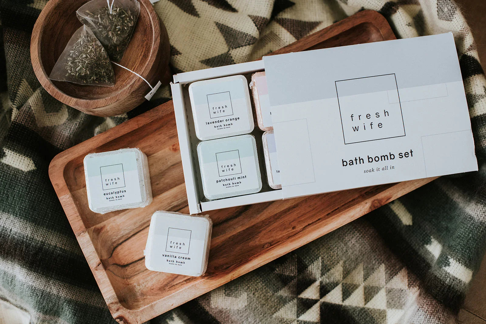 Fresh Wife bath bomb set on wooden tray with herbal tea bags and cozy blanket background