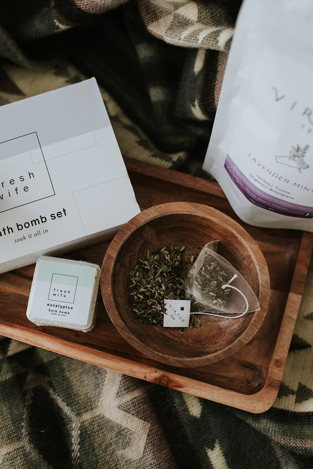 Cozy blanket with bath bomb set, herbal tea bag, and loose leaf tea on wooden tray