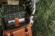 Open brown leather backpack with laptop inside, person wearing patterned blanket outdoors on grass