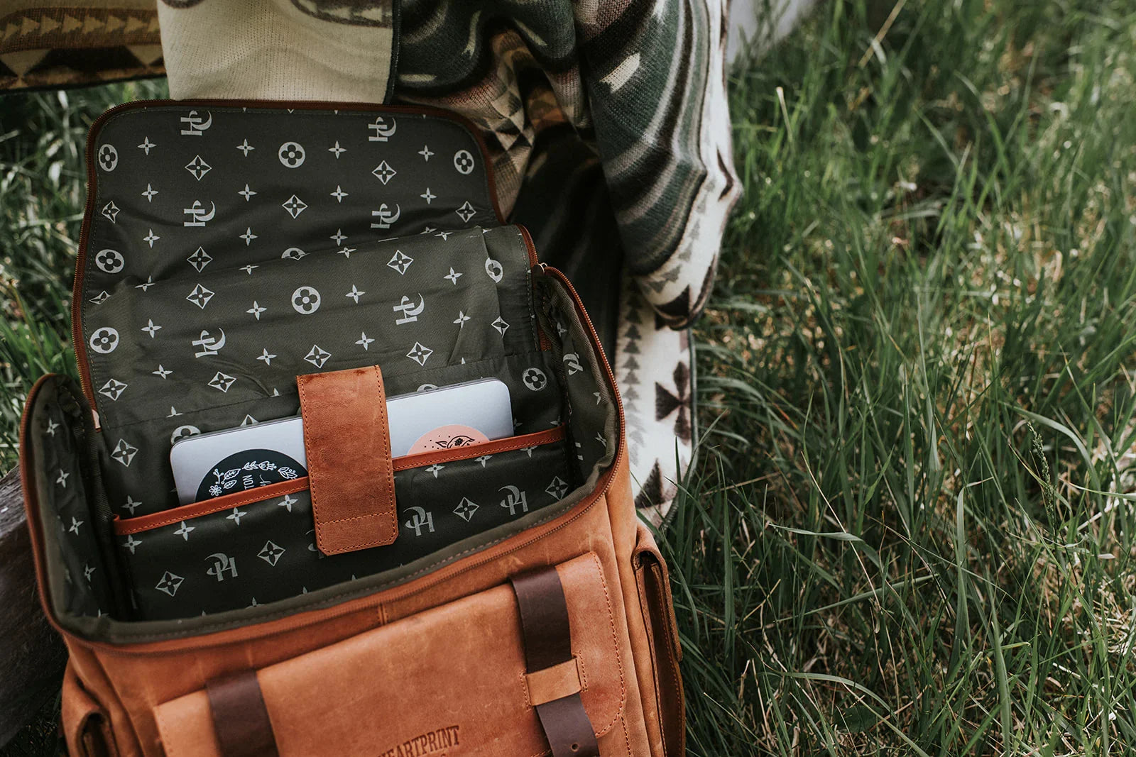 Open brown leather backpack with laptop inside, person wearing patterned blanket outdoors on grass