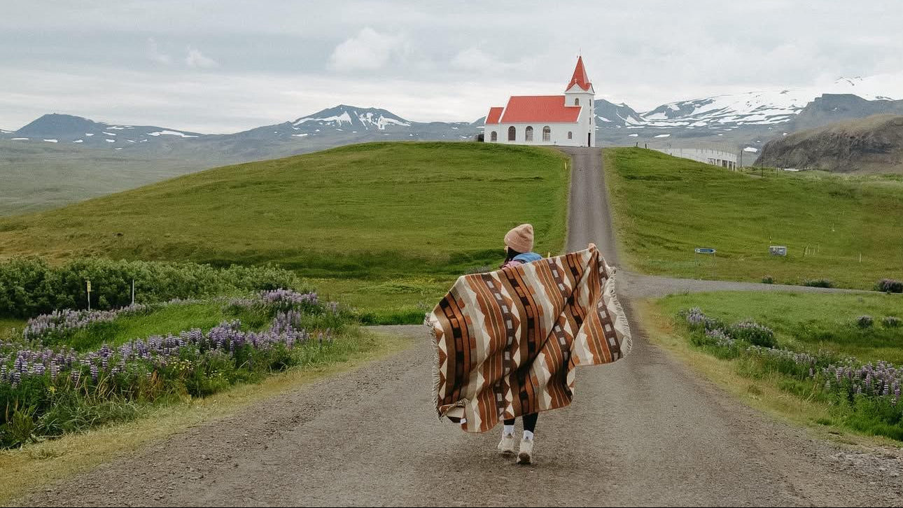 A person carrying a large brown and beige patterned blanket or wrap walks down a dirt road toward a small white church with a red roof on a grassy hill, with mountains and an overcast sky in the background.