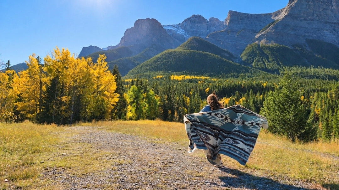 A person running outdoors in a mountain landscape, carrying or wrapped in a patterned blanket. The scene includes a gravel path, yellow autumn trees, evergreen forest, and rocky mountains under a bright blue sky with the sun shining.