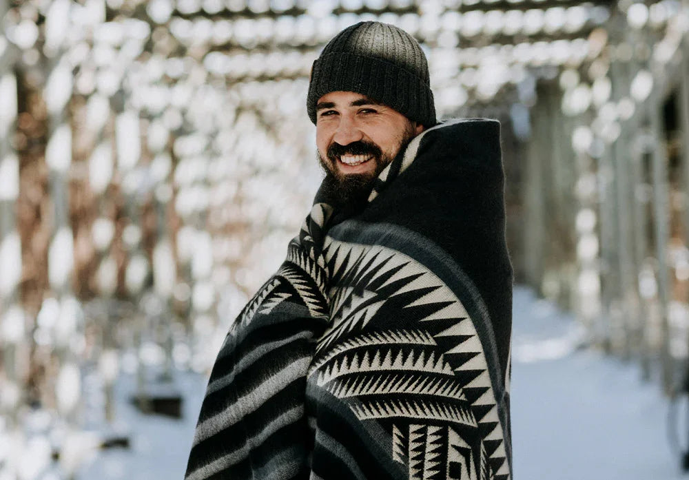 Man in knit beanie wrapped in cozy geometric patterned blanket outdoors in snowy setting