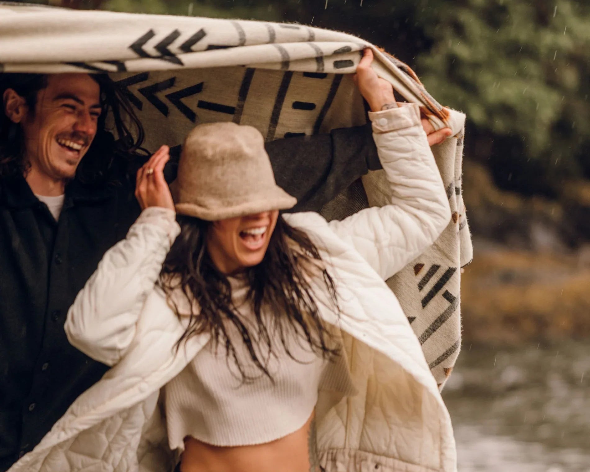 Two people walking together with a blanket over their heads near a body of water.