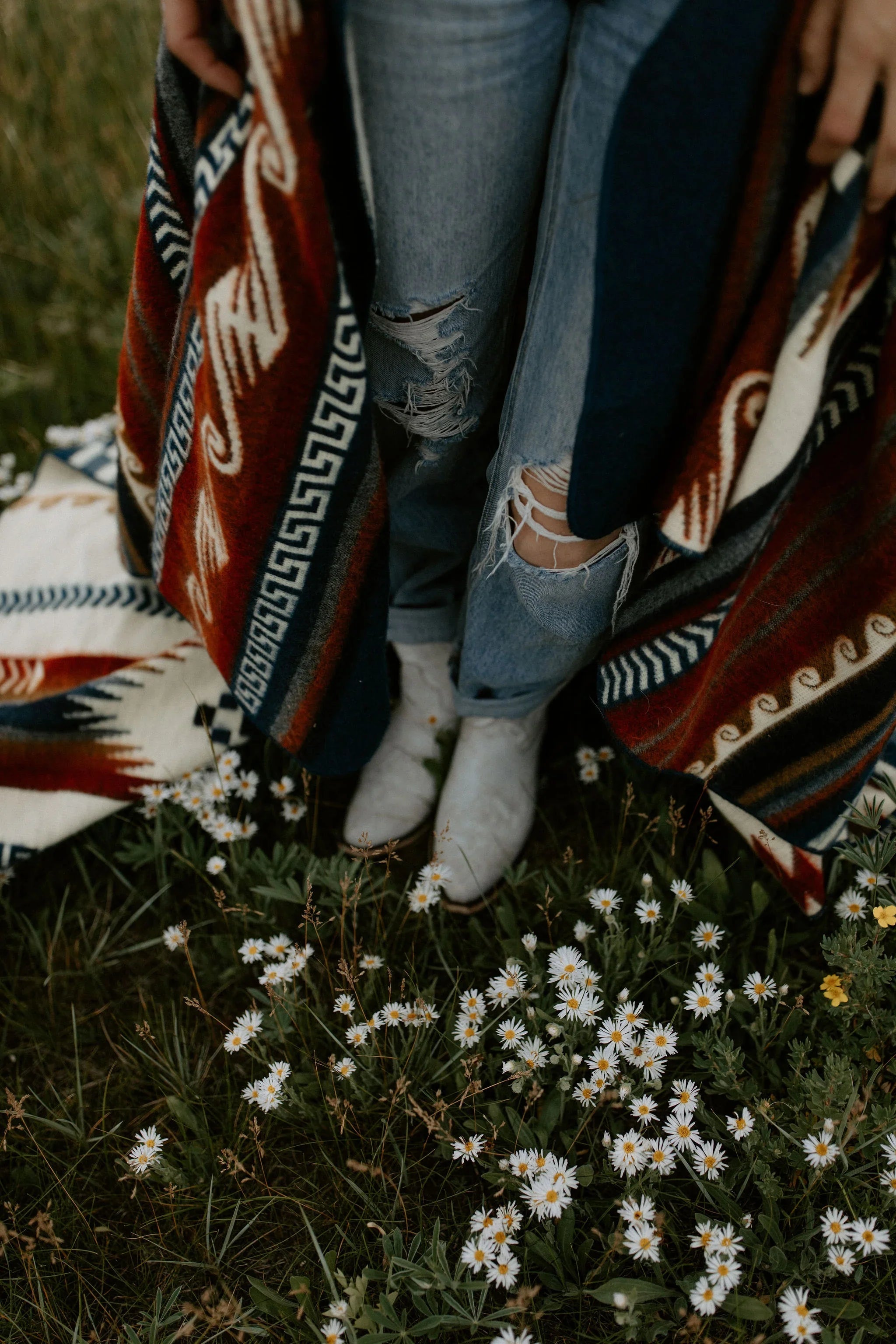 Person in ripped jeans and white boots wearing patterned blanket, standing in wildflowers