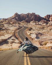 Woman wearing hat walking on desert road with geometric patterned blanket