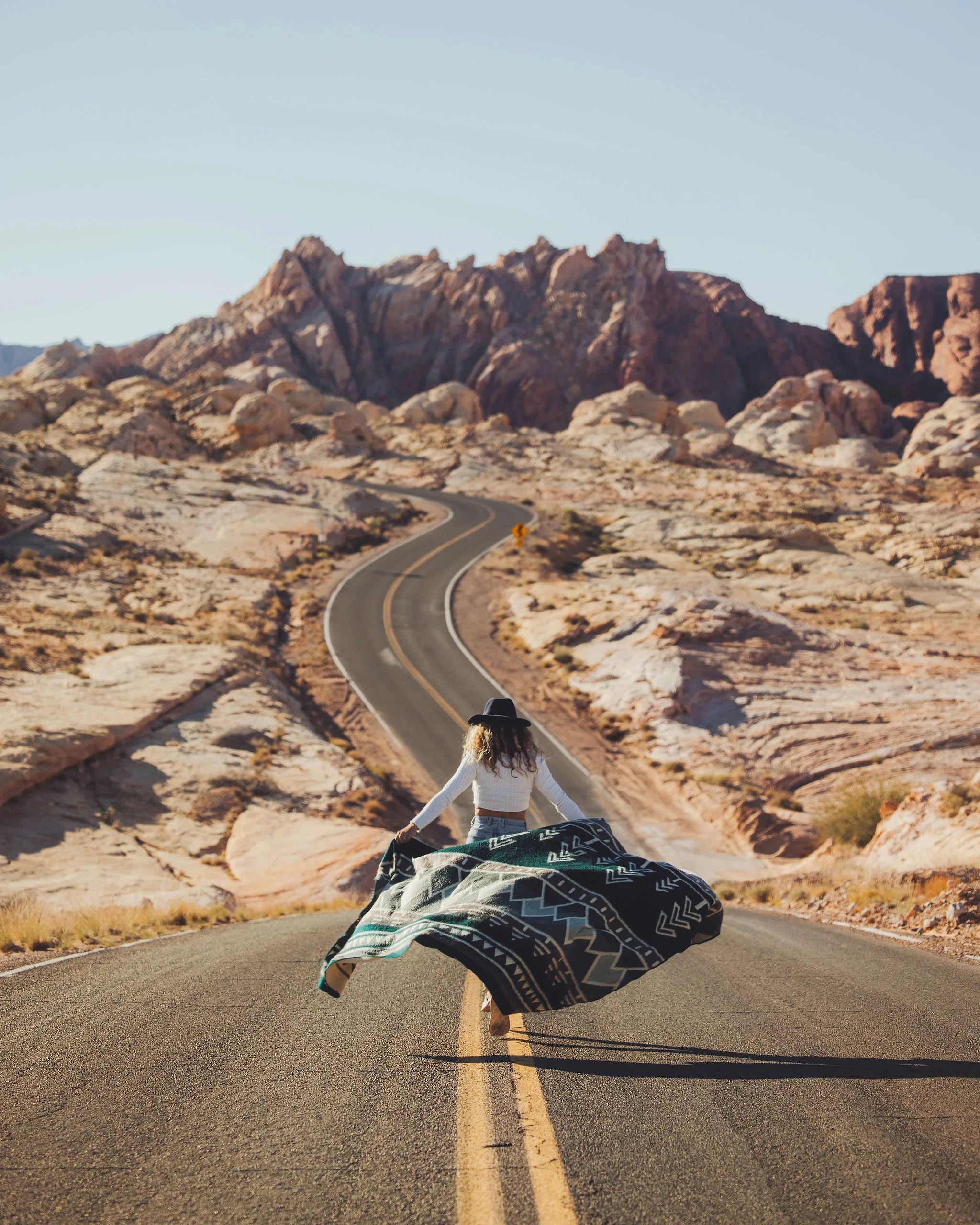 Woman wearing hat walking on desert road with geometric patterned blanket