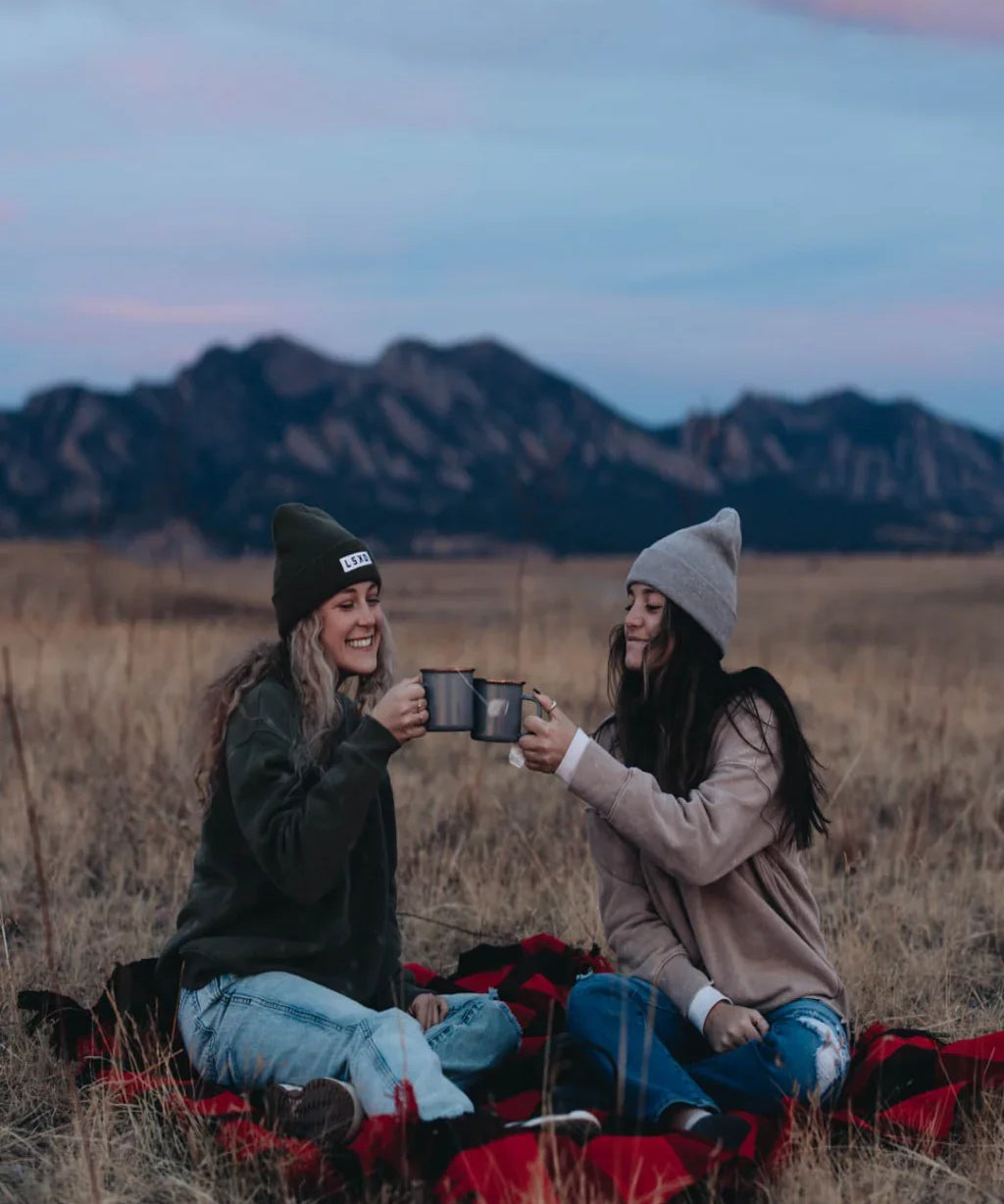 Two women on a plaid blanket outdoors, enjoying hot drinks with mountains at sunset