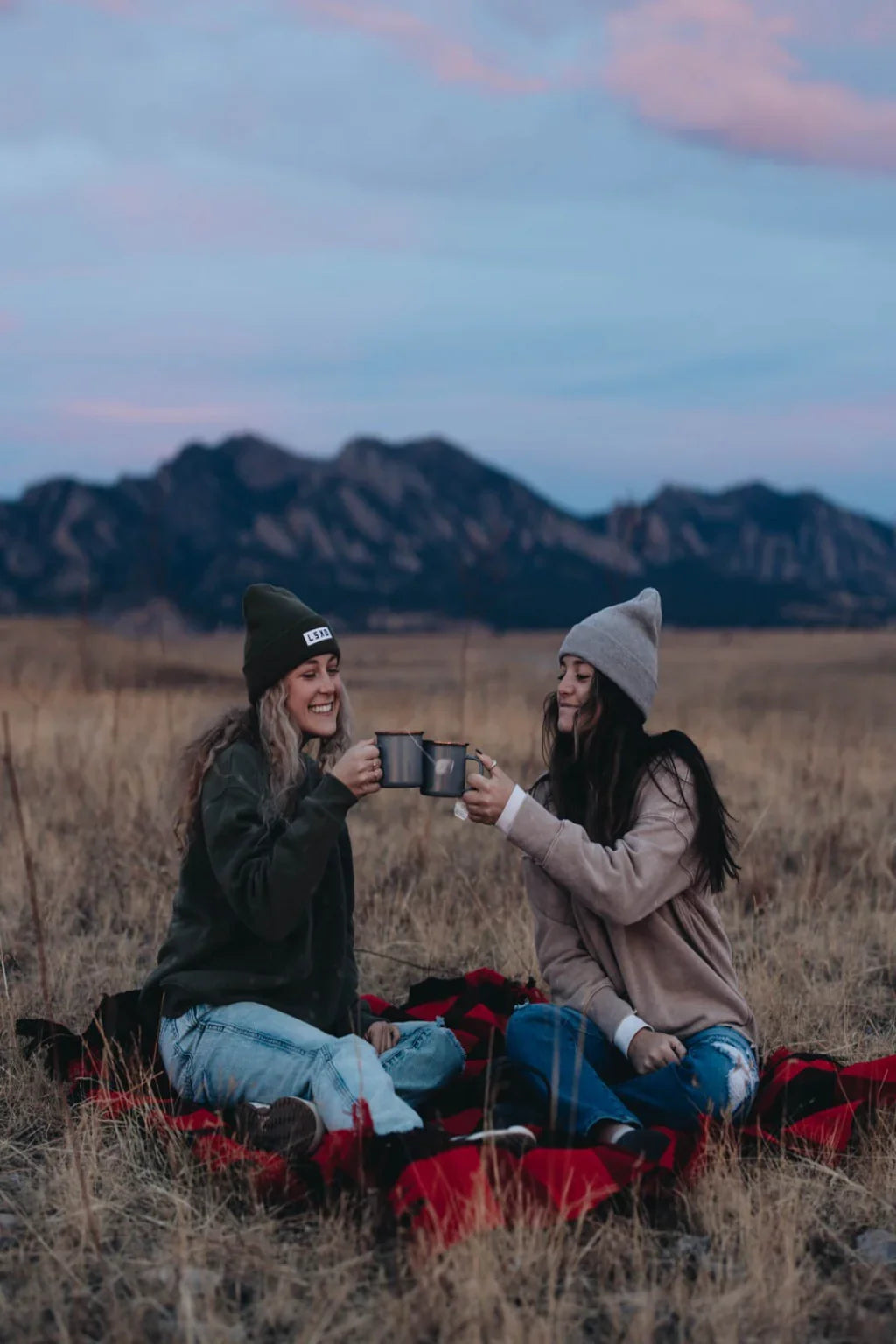 Two women on a plaid blanket outdoors, enjoying hot drinks with mountains at sunset