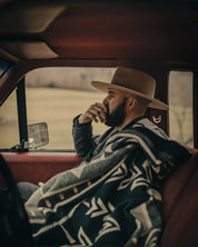 Man in wide-brim hat wrapped in patterned blanket sits in vintage truck, countryside view