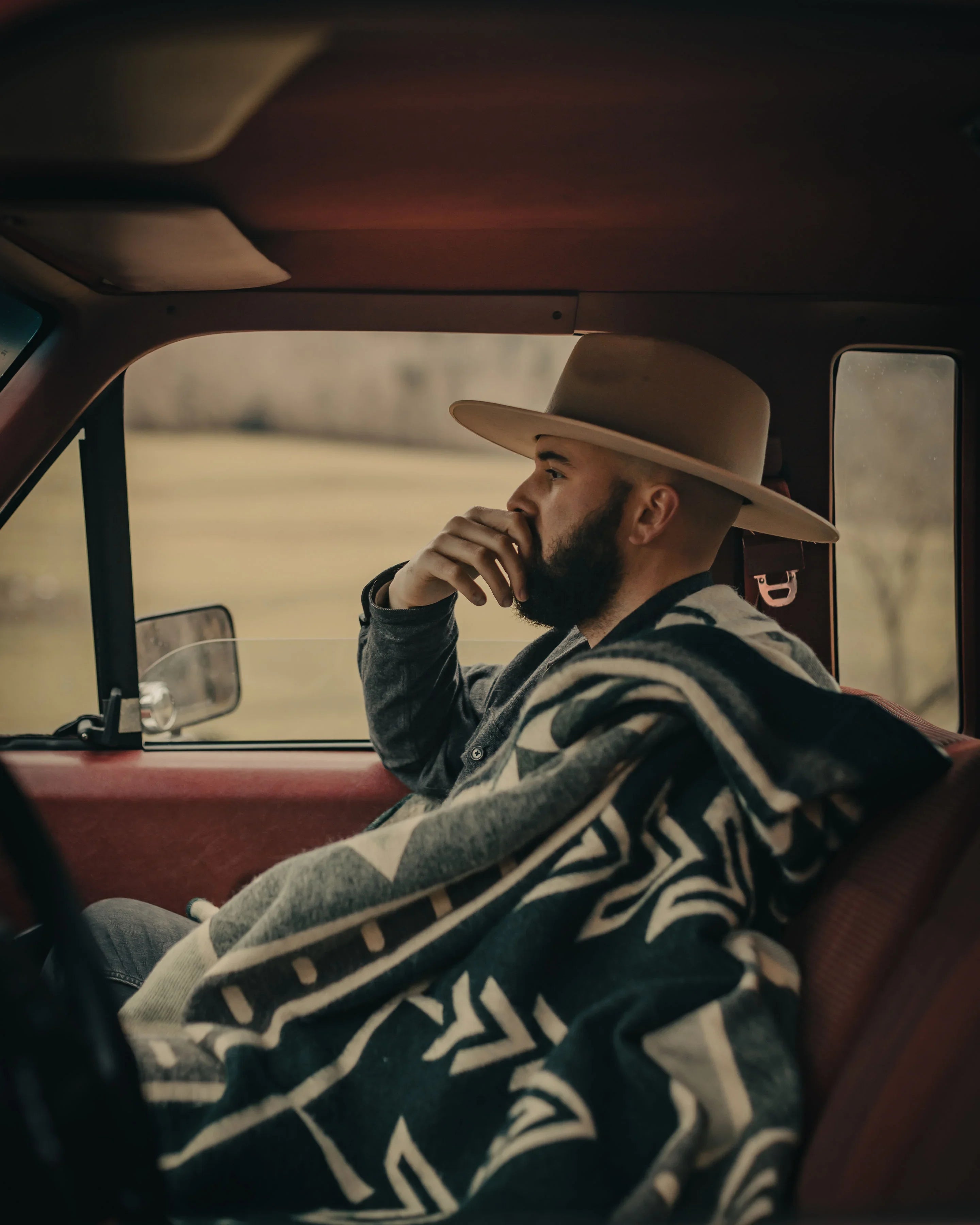 Man in wide-brim hat wrapped in patterned blanket sits in vintage truck, countryside view