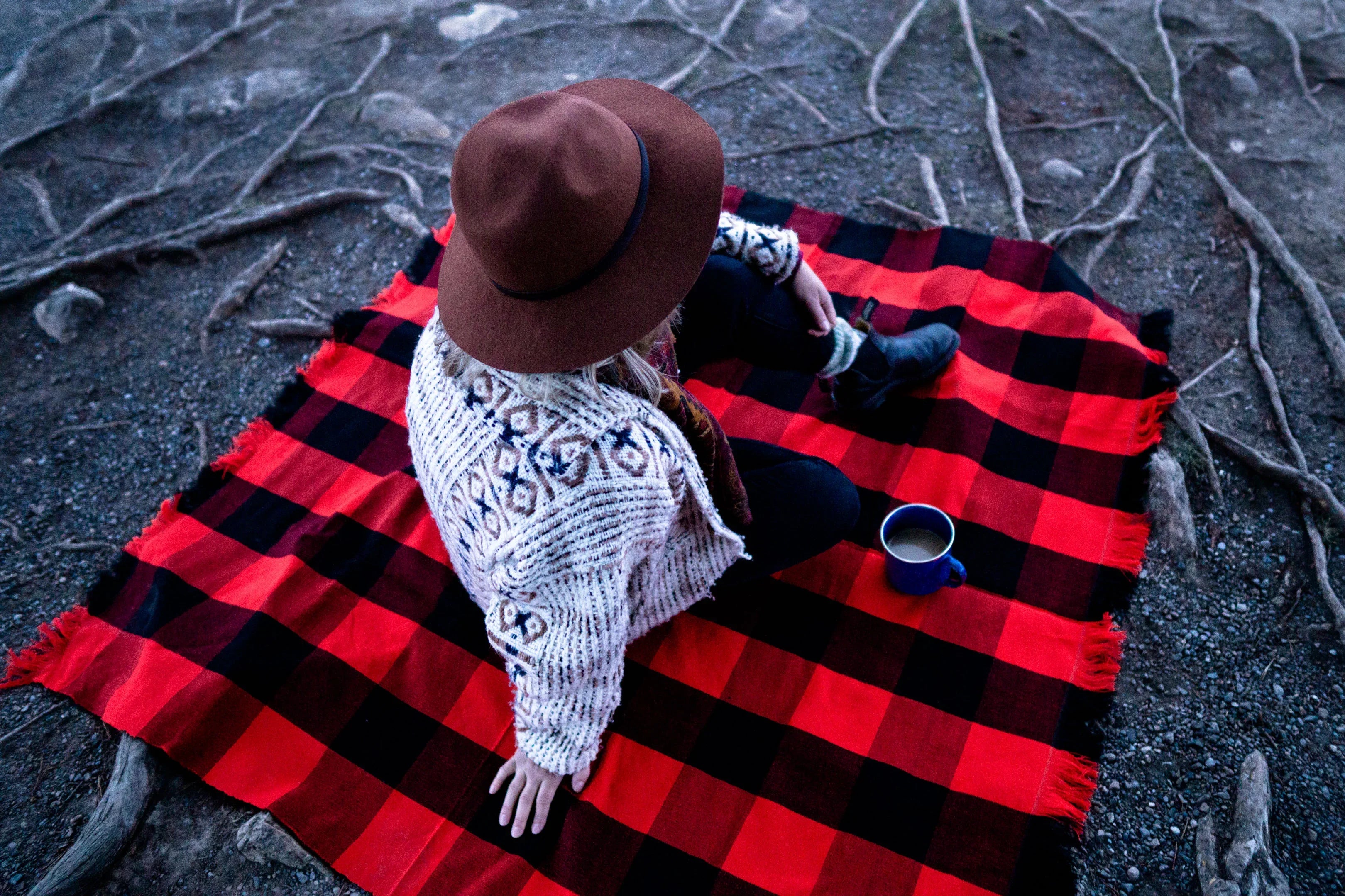Person sitting outdoors on a red and black buffalo plaid blanket with a coffee mug