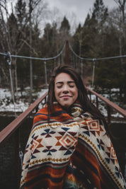 Woman wrapped in a patterned blanket on a rustic bridge in a winter forest
