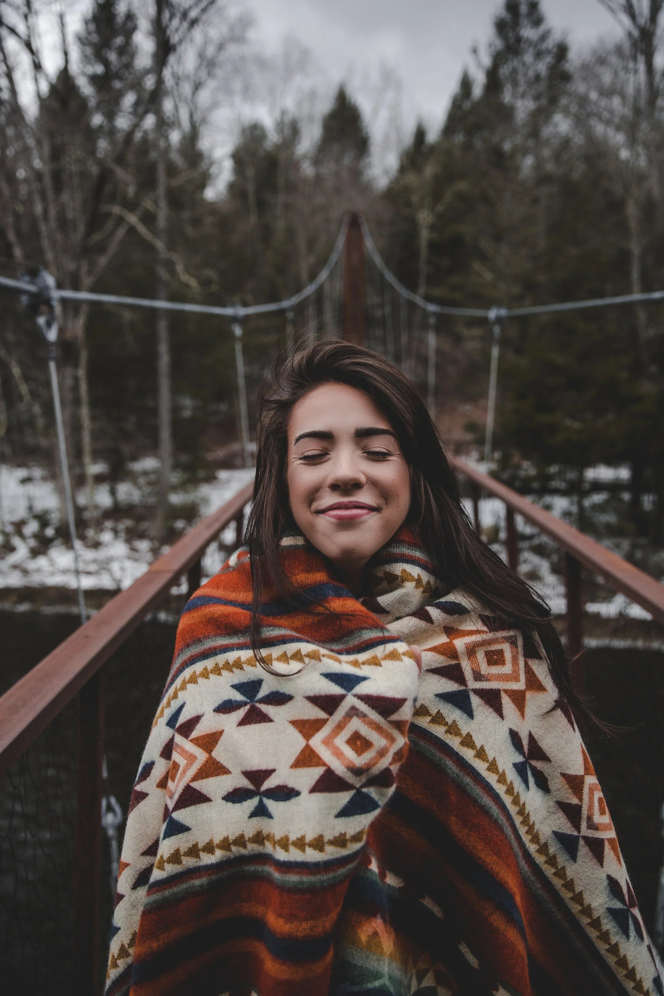 Woman wrapped in a patterned blanket on a rustic bridge in a winter forest