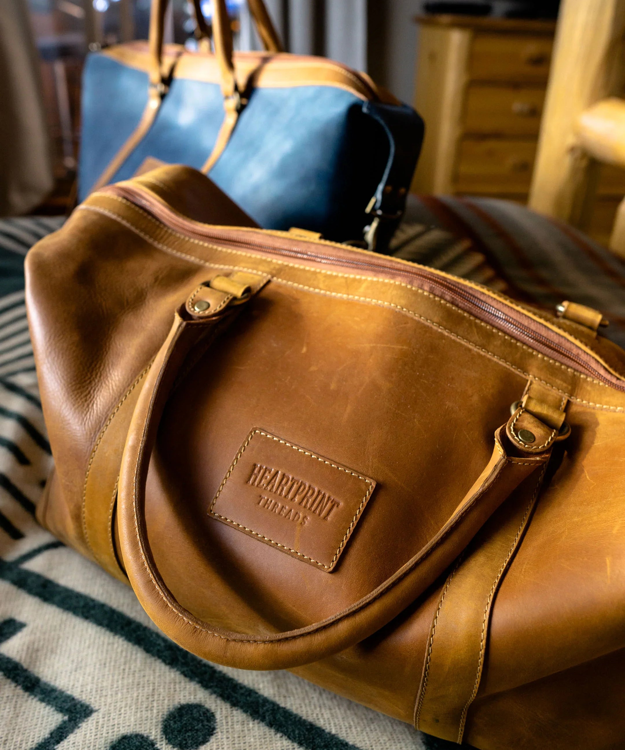Brown leather duffel bag labeled Heartprint Threads on patterned blanket, blue duffel in background