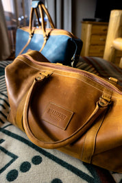 Brown leather duffel bag labeled Heartprint Threads on patterned blanket, blue duffel in background