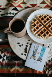 Breakfast scene with coffee, waffles, and love card on patterned cozy blanket.