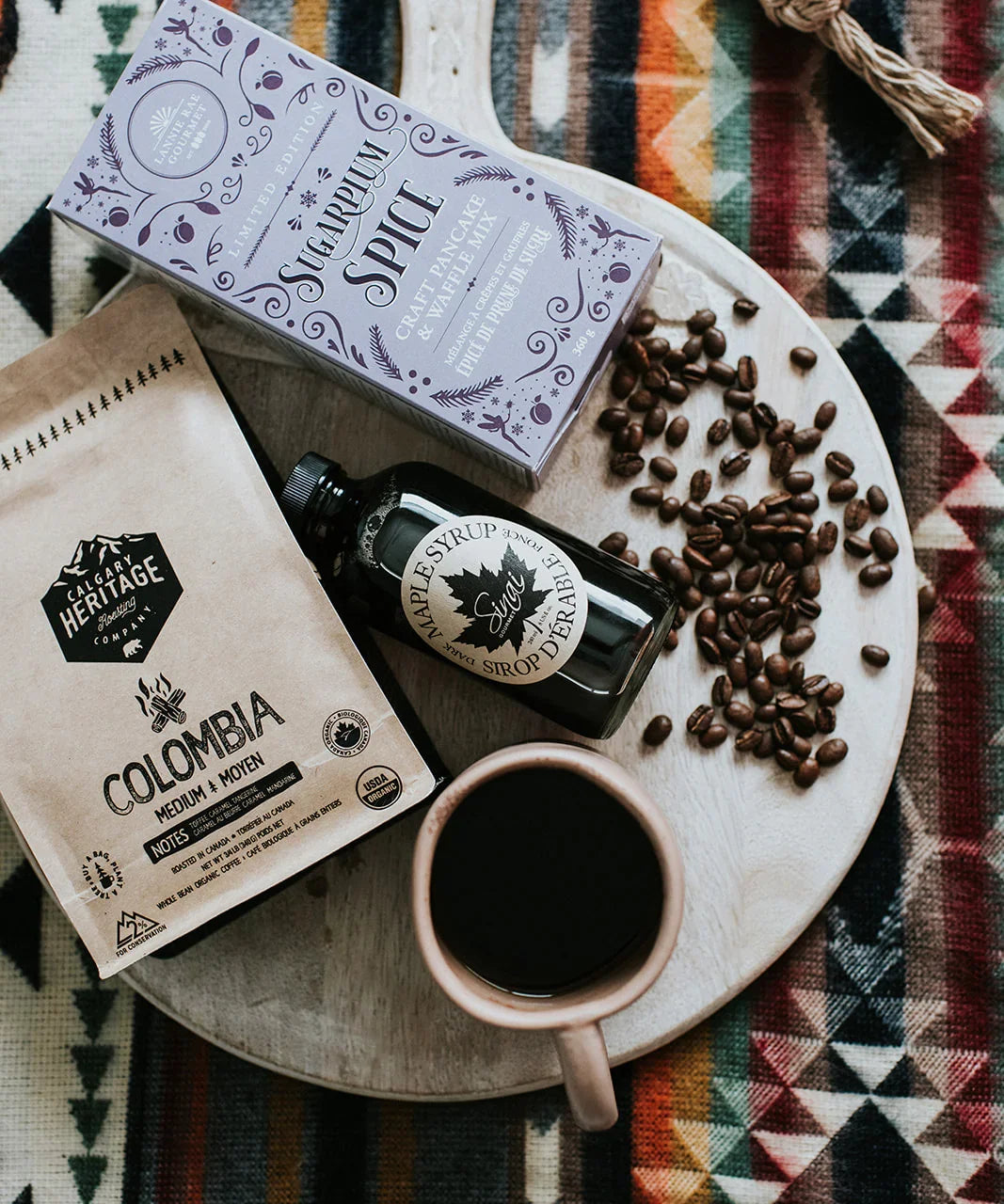 Coffee beans, Colombian coffee, maple syrup, and spiced pancake mix on a serving board over a patterned blanket