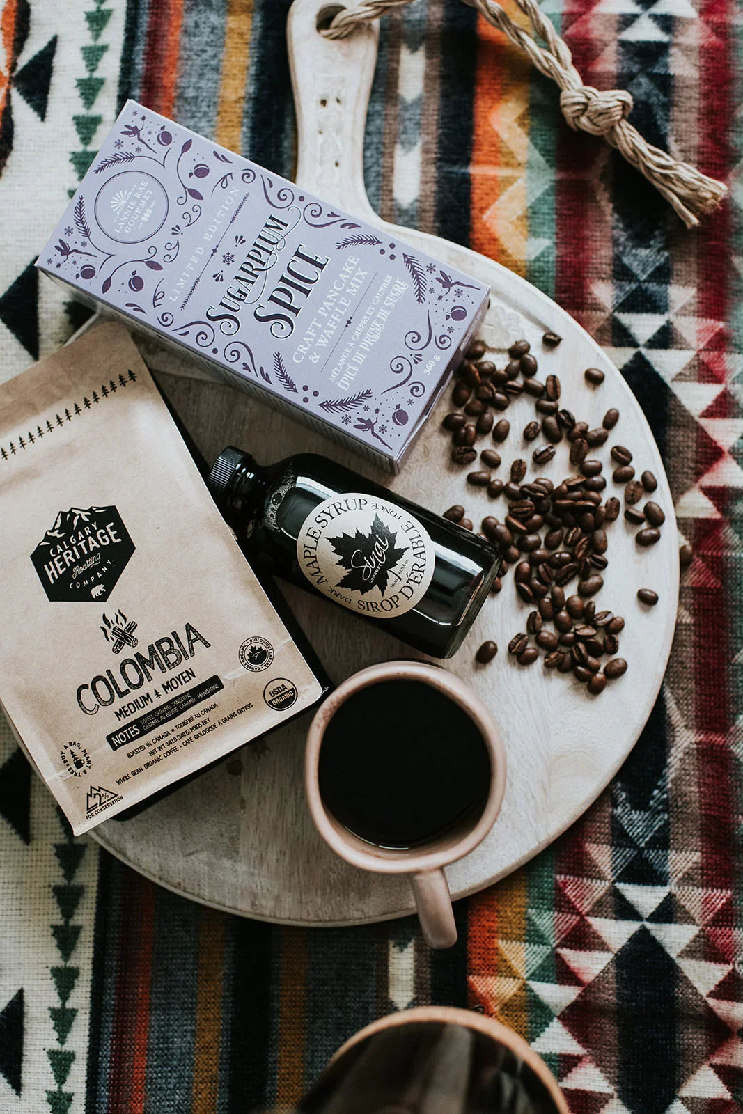 Coffee beans, Colombian coffee, maple syrup, and spiced pancake mix on a serving board over a patterned blanket