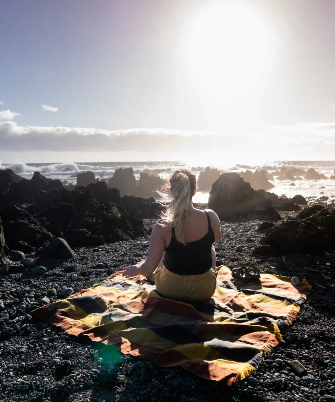 Woman sitting on a plaid blanket by rocky seashore under bright sunlight