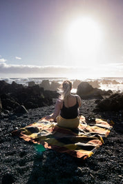 Woman sitting on a plaid blanket by rocky seashore under bright sunlight