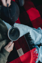 Close-up of person pouring tea over red and black plaid fringed blanket on lap