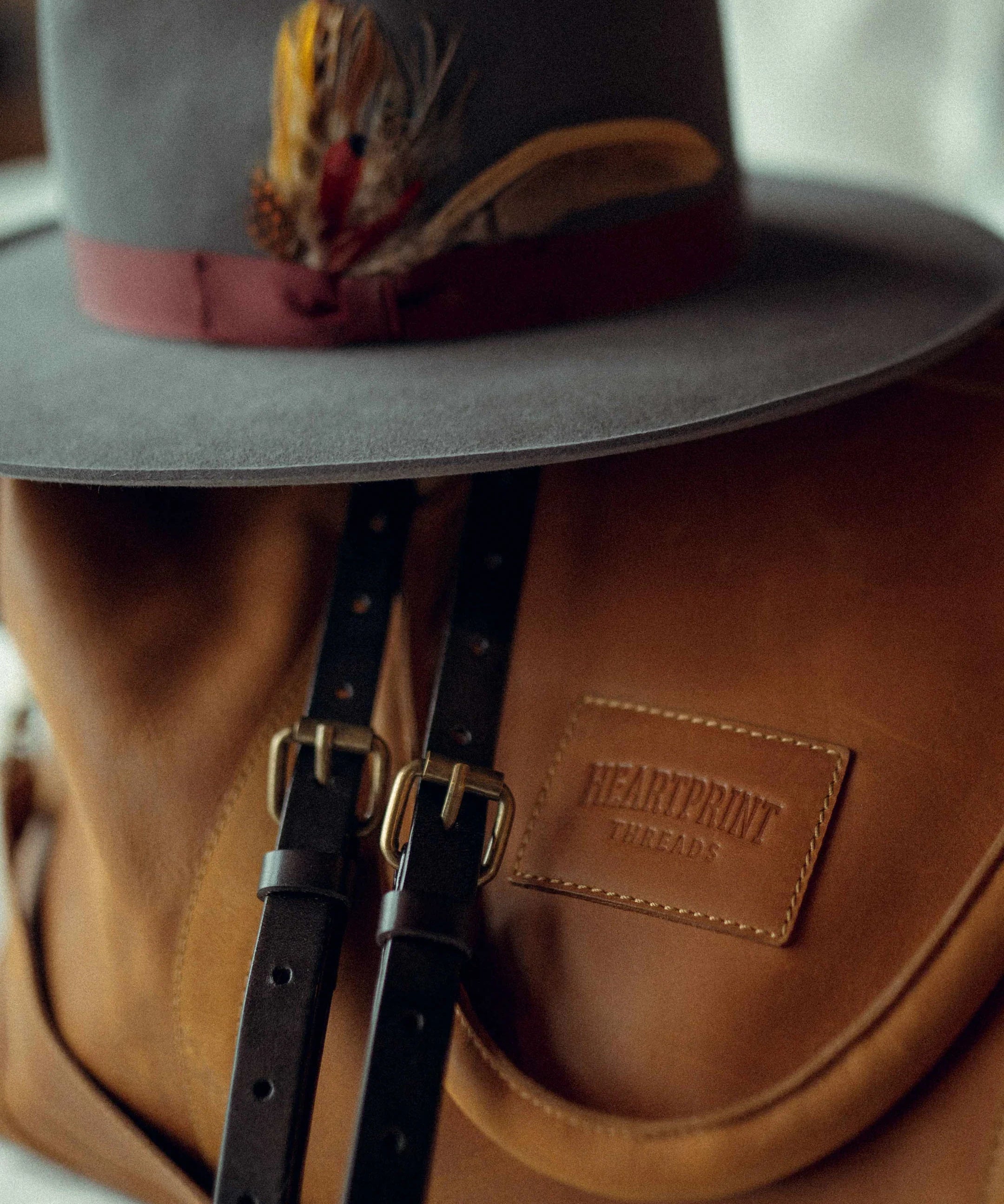 Grey felt hat with feather detail resting on brown leather backpack with buckle straps
