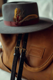 Grey felt hat with feather detail resting on brown leather backpack with buckle straps