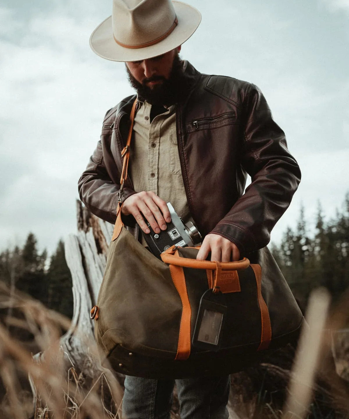 Man in hat and leather jacket outdoors holding a green canvas duffel bag with brown straps