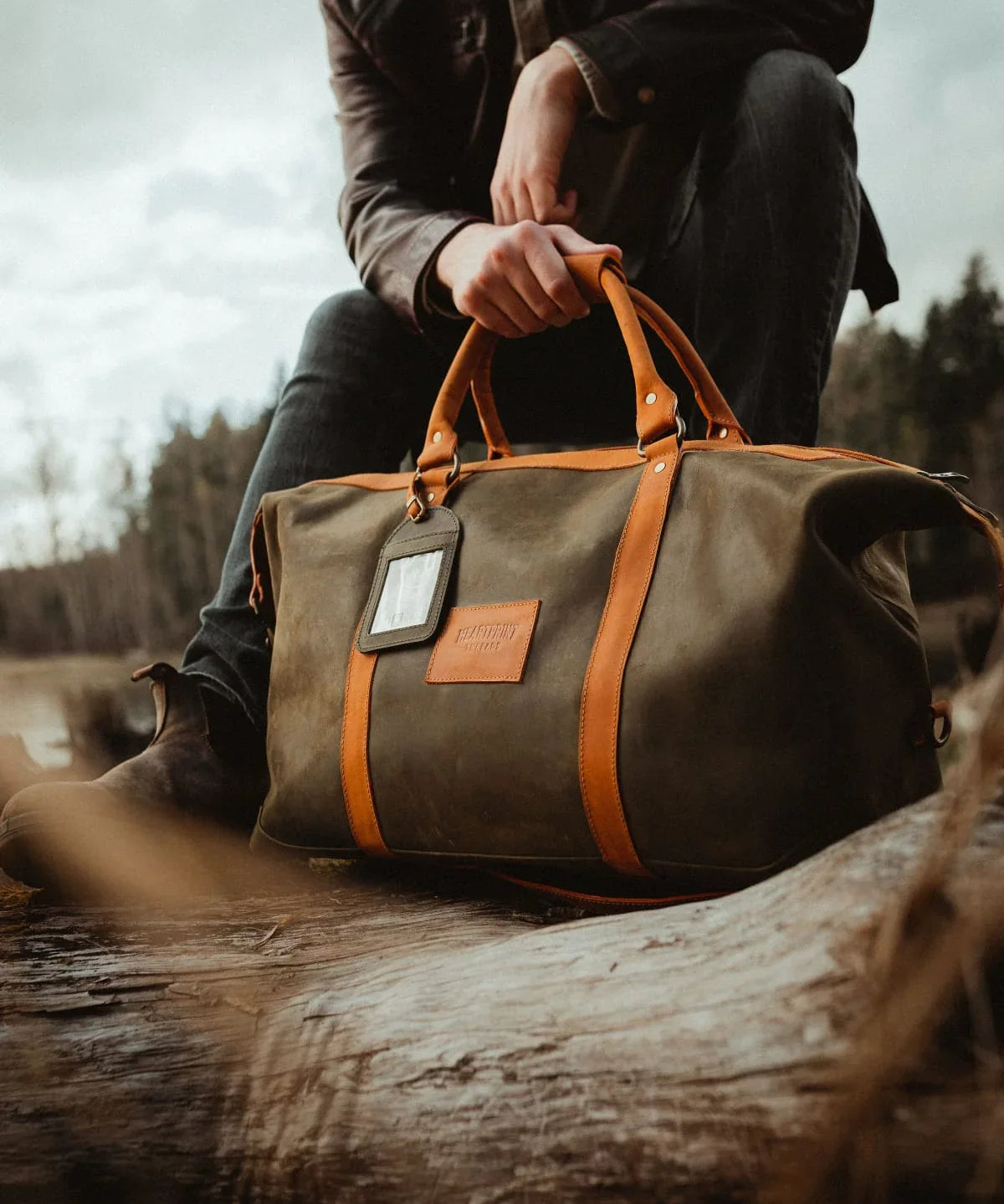 Man outdoors with olive green and tan leather duffle bag on a log