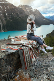 Woman sitting on patterned blanket atop rock ledge overlooking lake and mountains