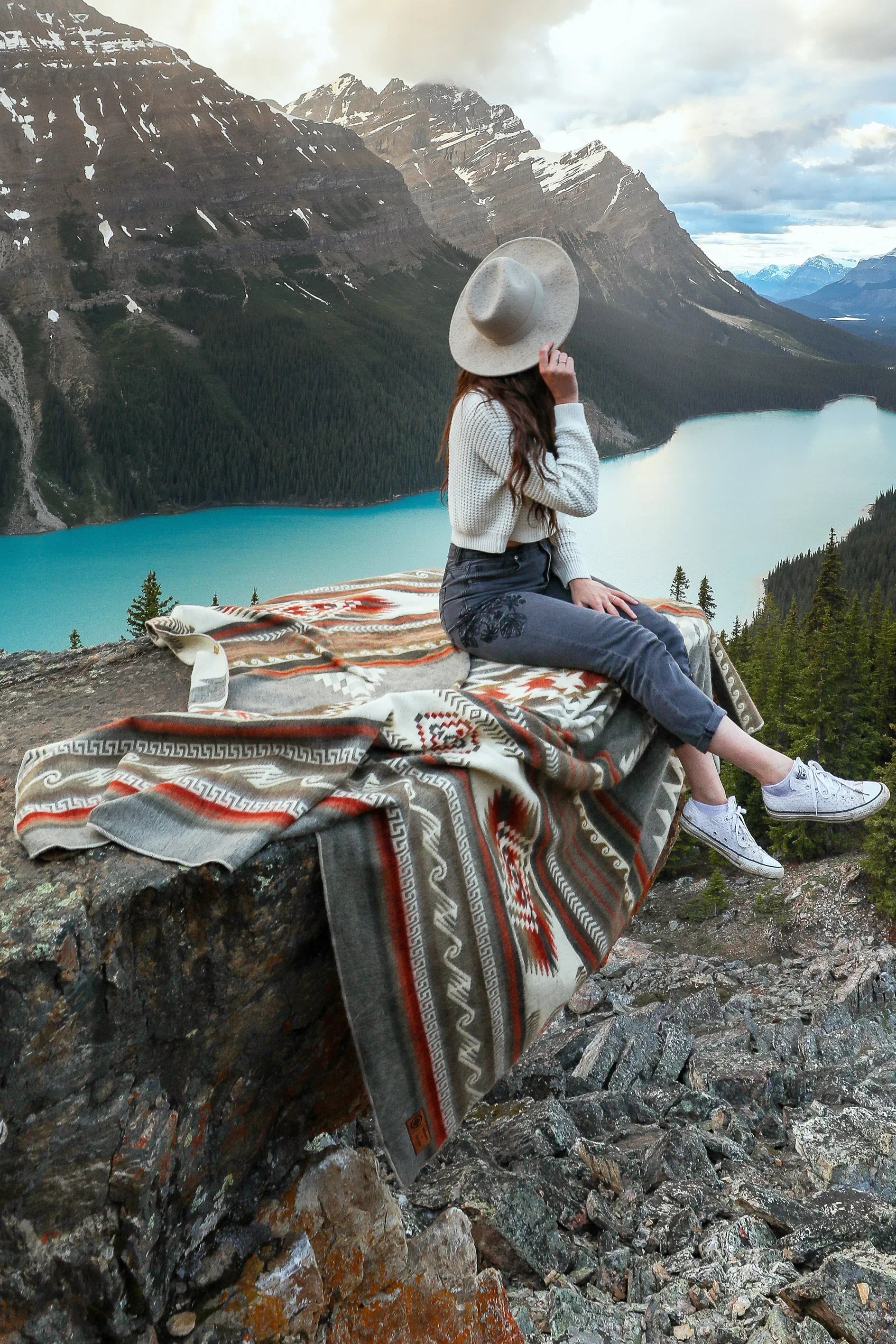 Woman sitting on patterned blanket atop rock ledge overlooking lake and mountains
